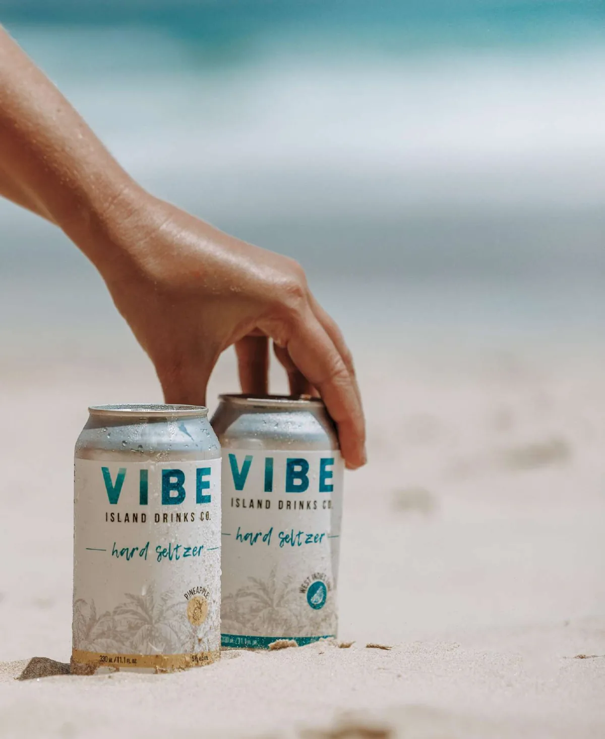 A hand placing two cans of Vibe Hard Seltzer in beach sand