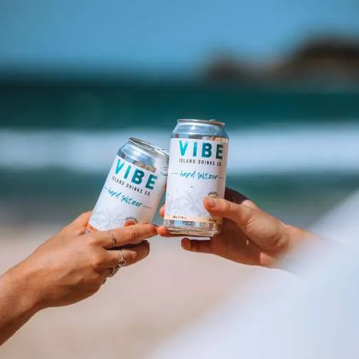 Two cans of Vibe raised in a toast over turquoise water
